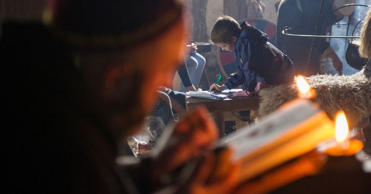 man dressed an Anglo-Saxon writing in a book by candle light with a young boy in background